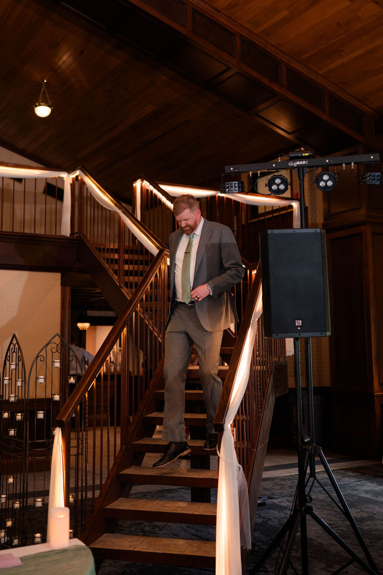 Groom in gray suit descends wooden staircase inside rustic wedding reception venue with warm lighting.
