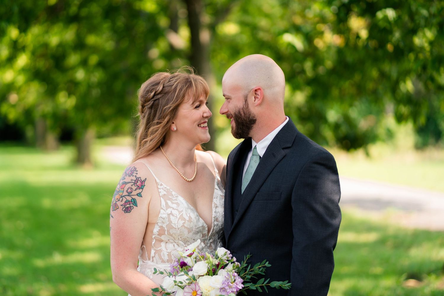 A smiling bride in a lace gown and tattooed arm holds a bouquet while gazing at her bald bearded groom.