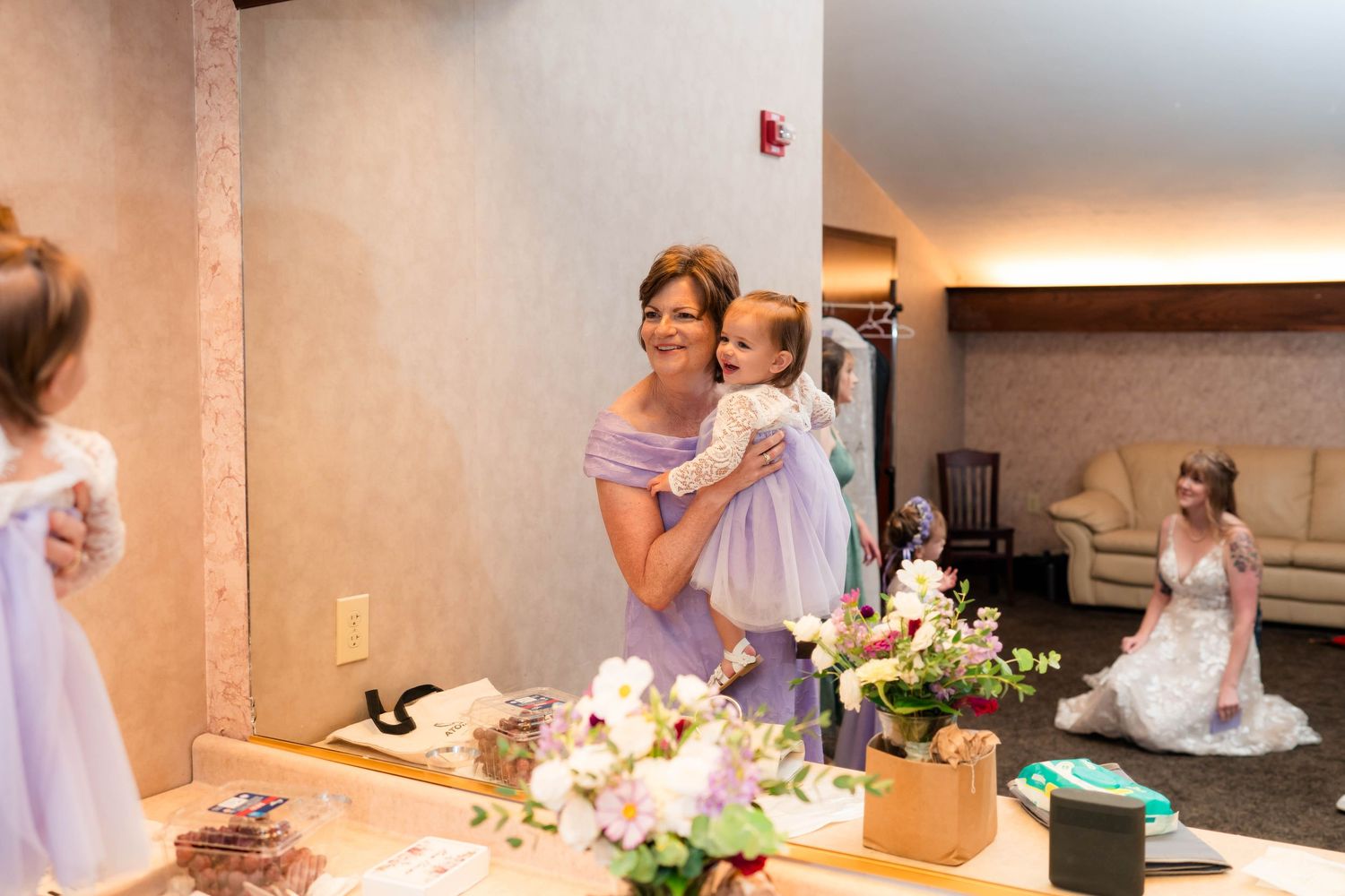 Woman in lavender dress holds young child in white dress at wedding venue with flowers.