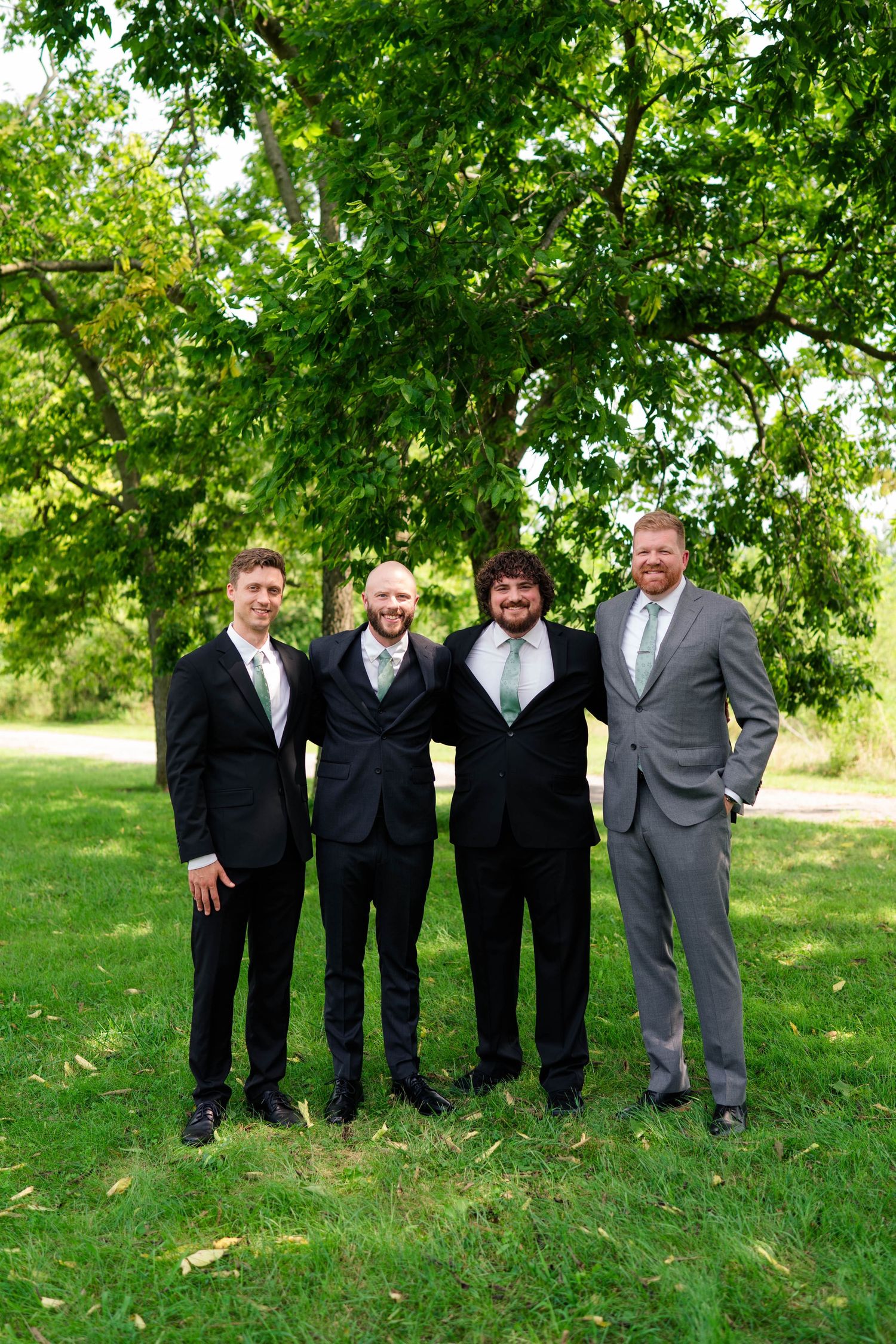 Four groomsmen in black and gray suits smile together under a leafy tree at an outdoor wedding celebration.