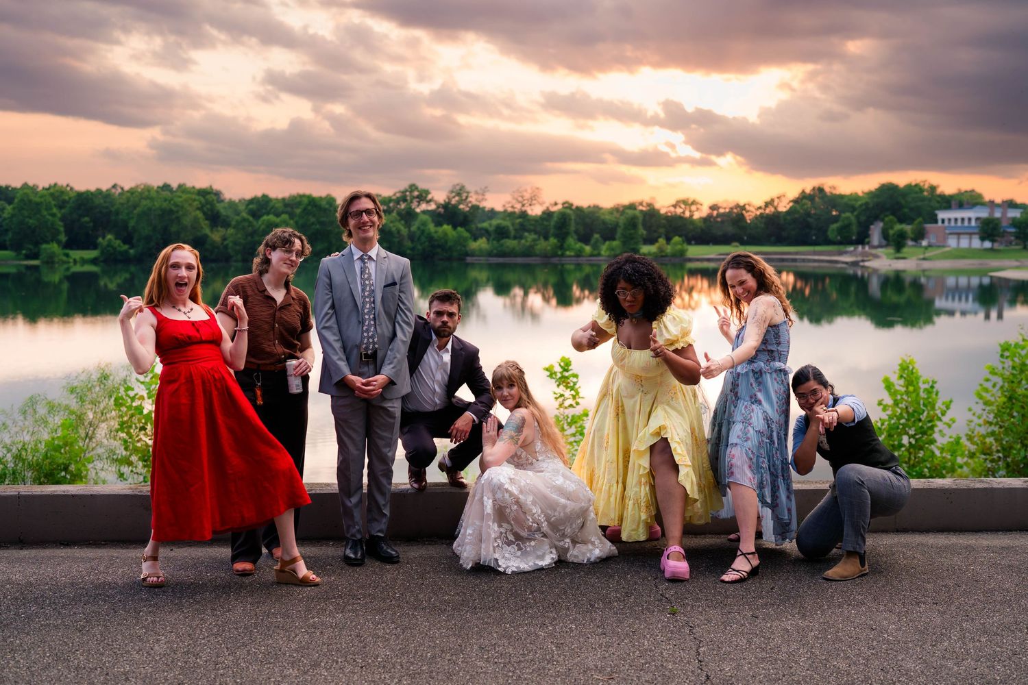 Wedding party poses dramatically by lake at sunset in colorful formal attire.