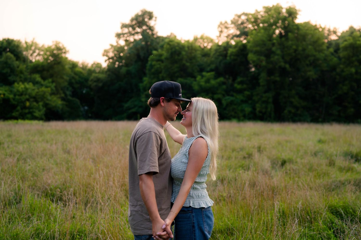 Young couple sharing a kiss at sunset in an open grassy field surrounded by trees.
