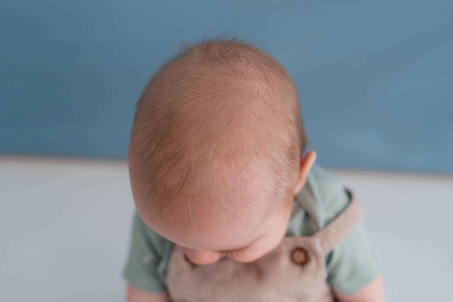Top view of a baby with fine hair wearing a sage green onesie and tan overalls against a blue background.