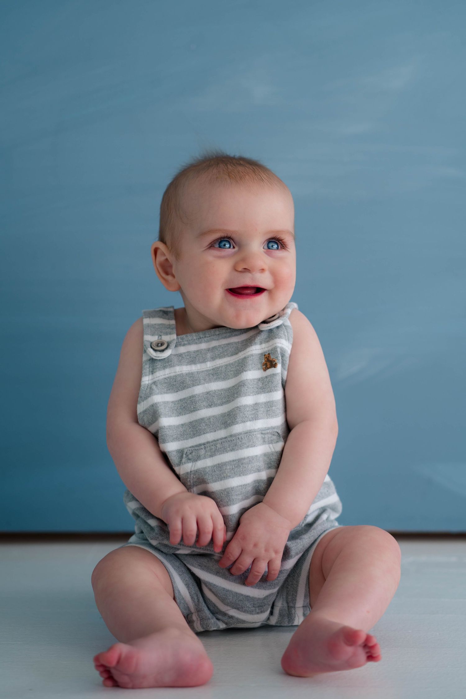 Happy blue-eyed baby in striped gray romper sitting and smiling against blue background.