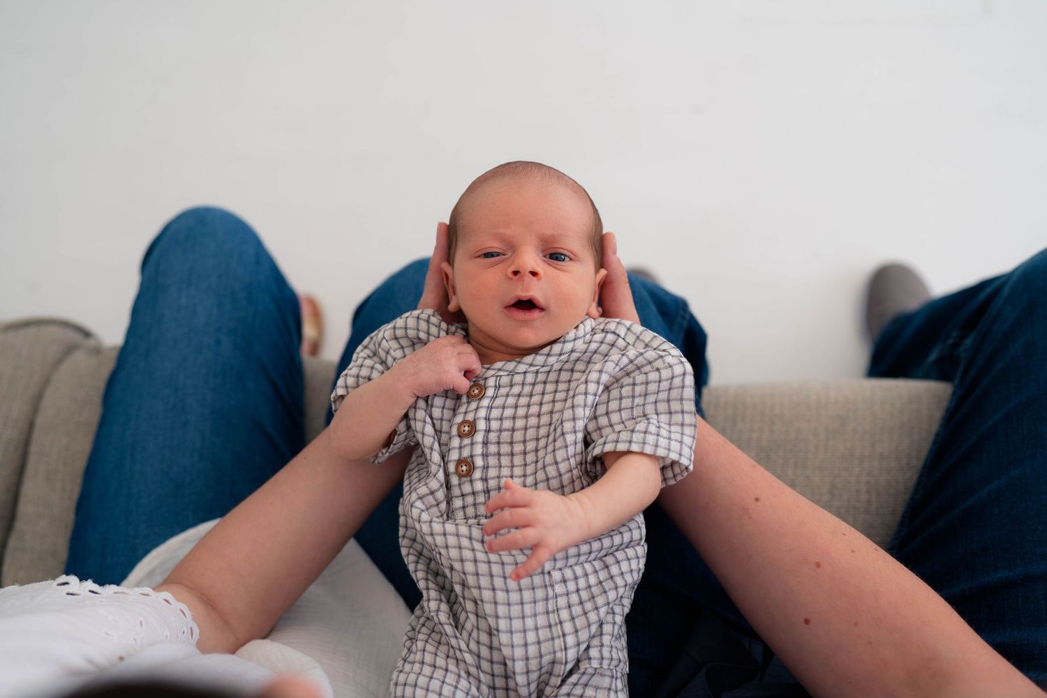 A newborn baby in a checkered onesie is gently supported by parents hands on a cozy couch.