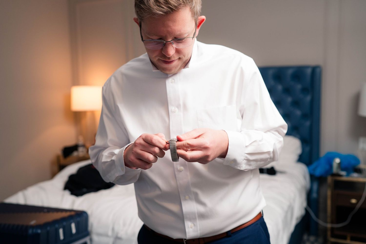 Groom in white dress shirt fastening cufflinks while getting ready in a hotel room.