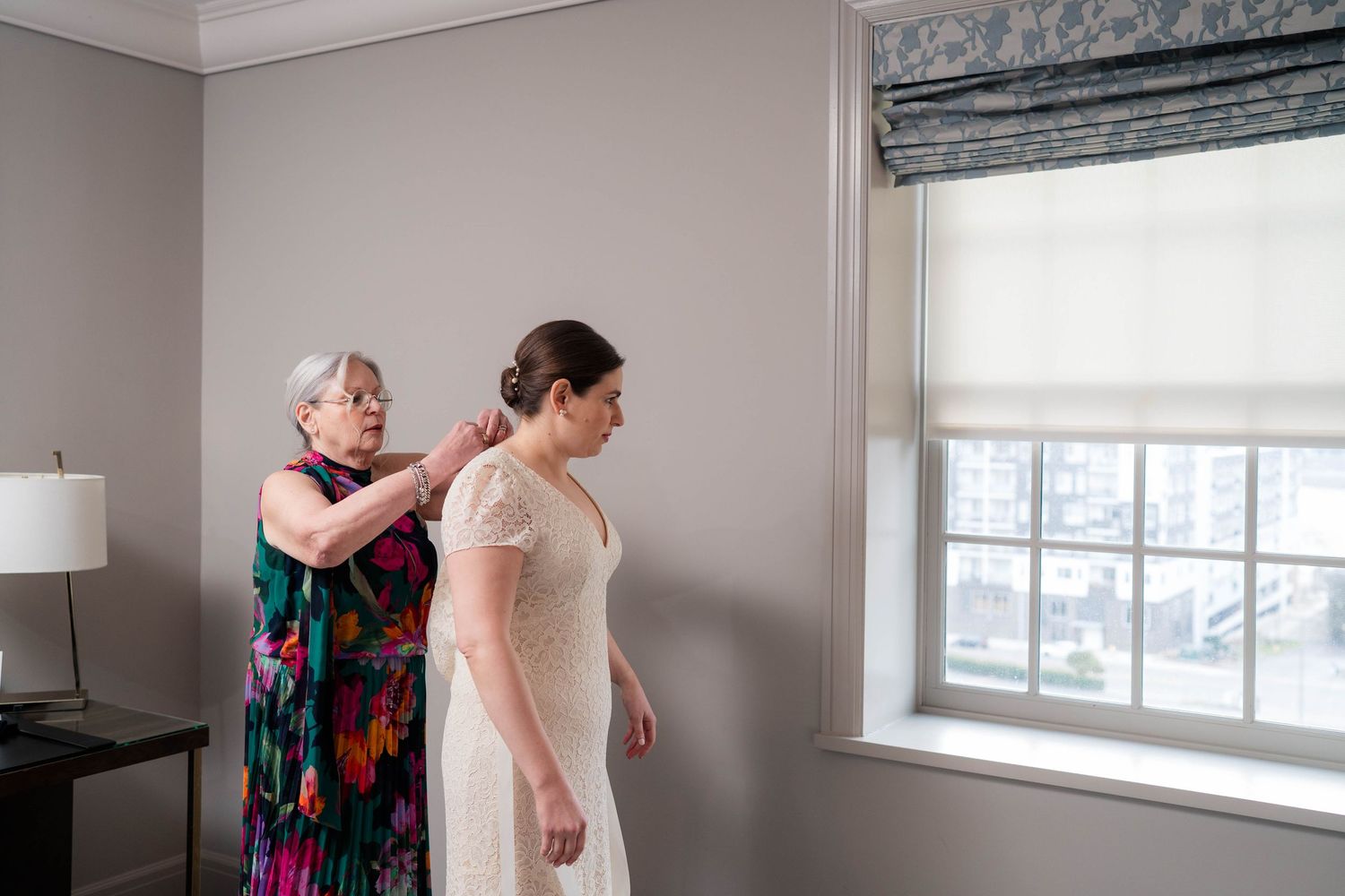 Older woman in floral dress helps bride with necklace near bright hotel window.
