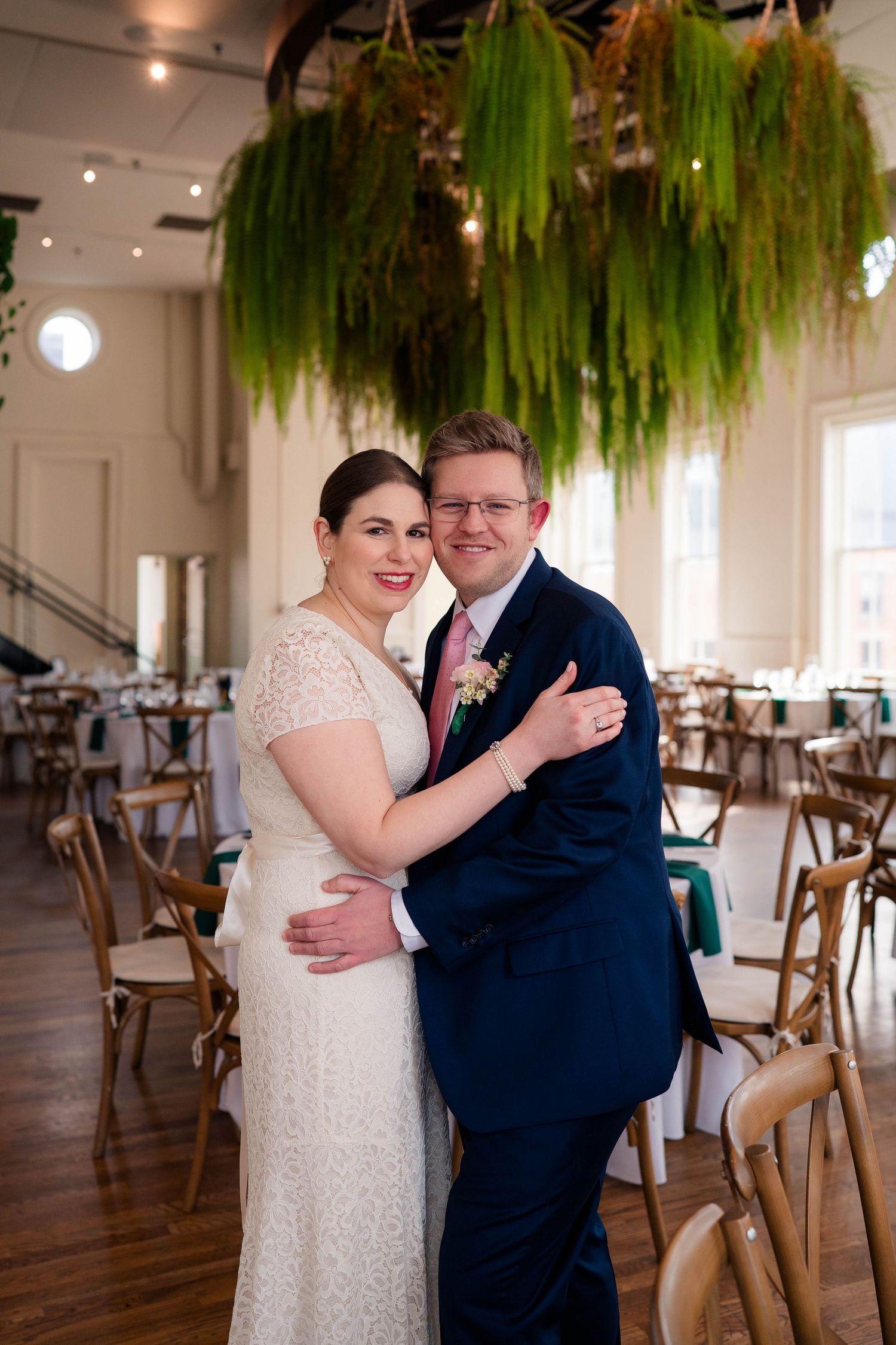 Bride and groom embracing and smiling beneath a stunning hanging greenery installation in a bright wedding venue.