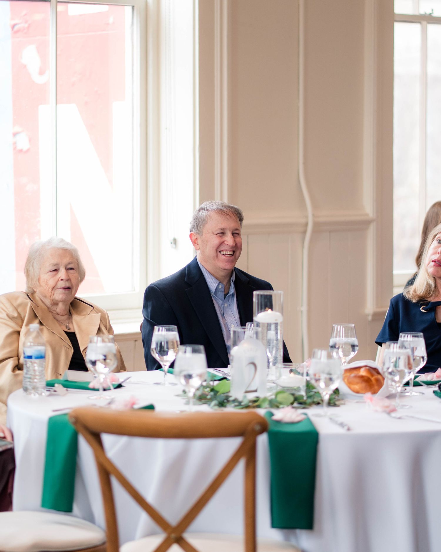 Three wedding guests smile and laugh together at a reception table near a bright window.