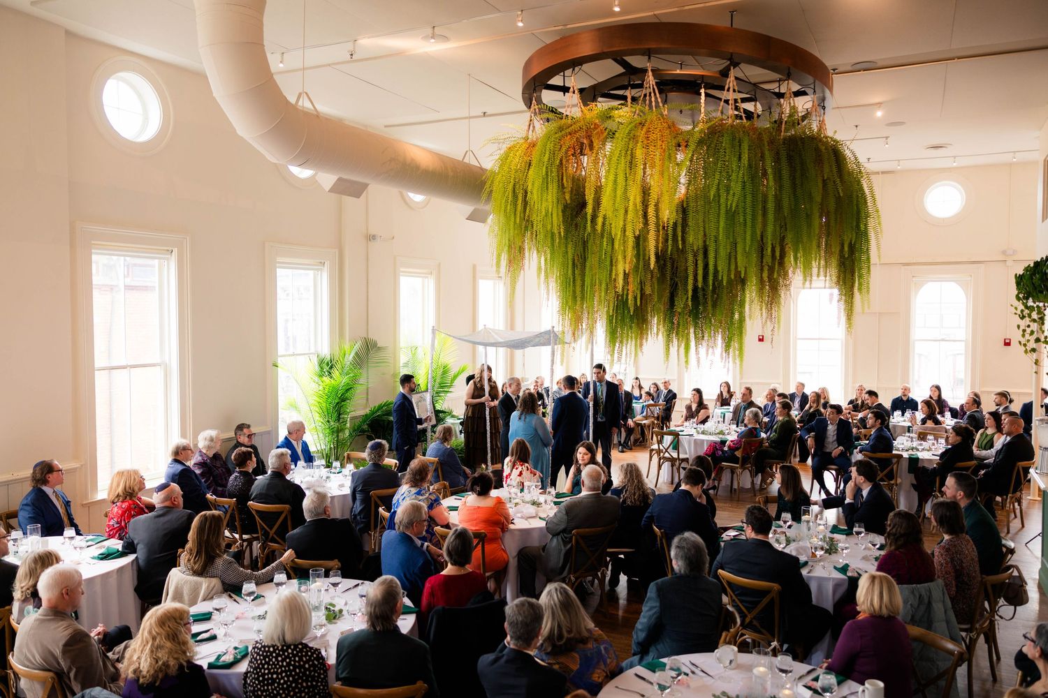 Elegant wedding reception in bright hall with hanging greenery centerpiece and seated guests.