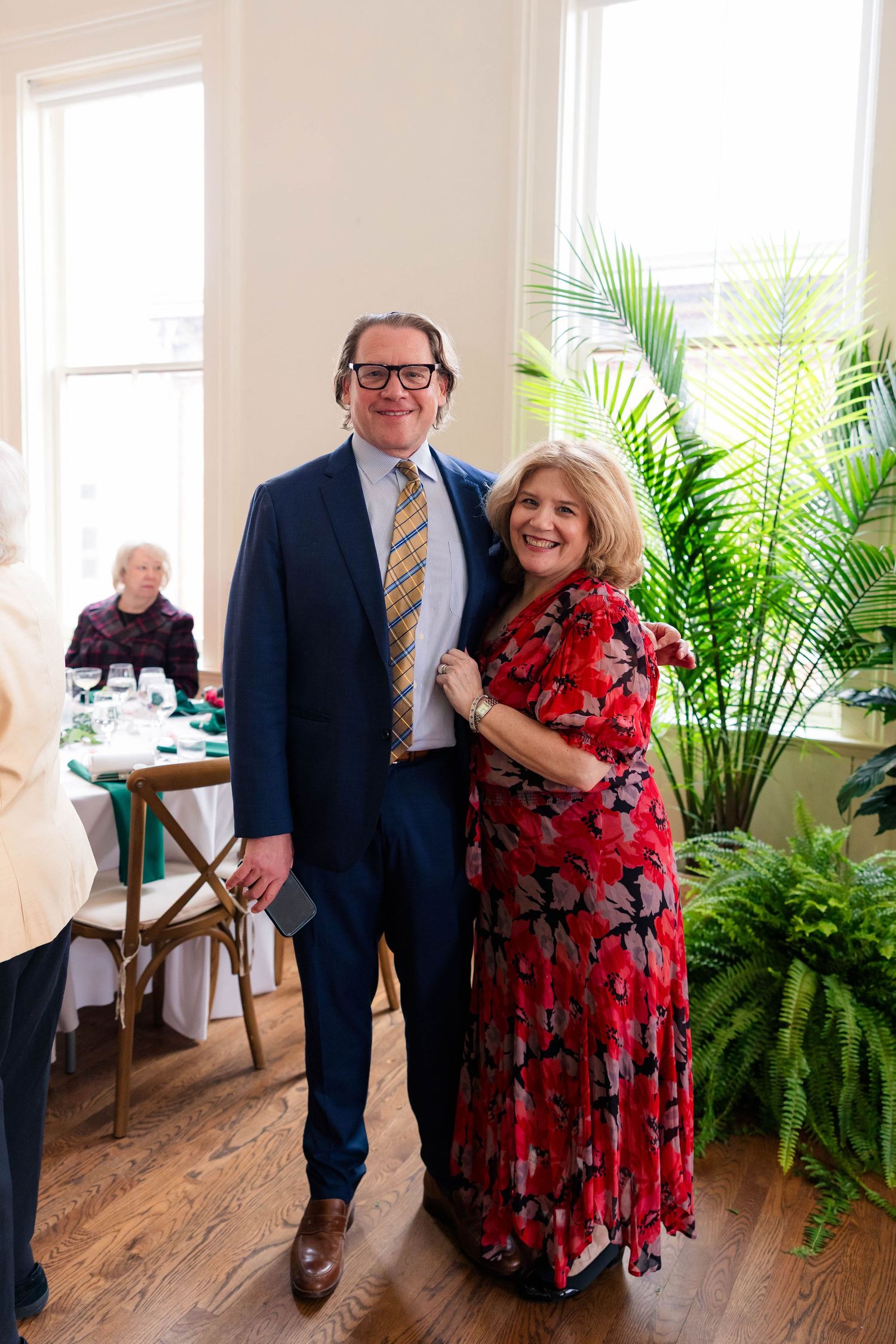 Couple posing together at elegant indoor event, man in suit and woman in red floral dress.