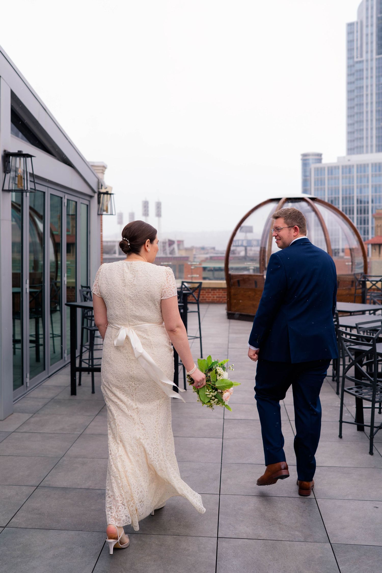 Bride and groom walk hand in hand on a rooftop terrace with a city skyline view on their wedding day.