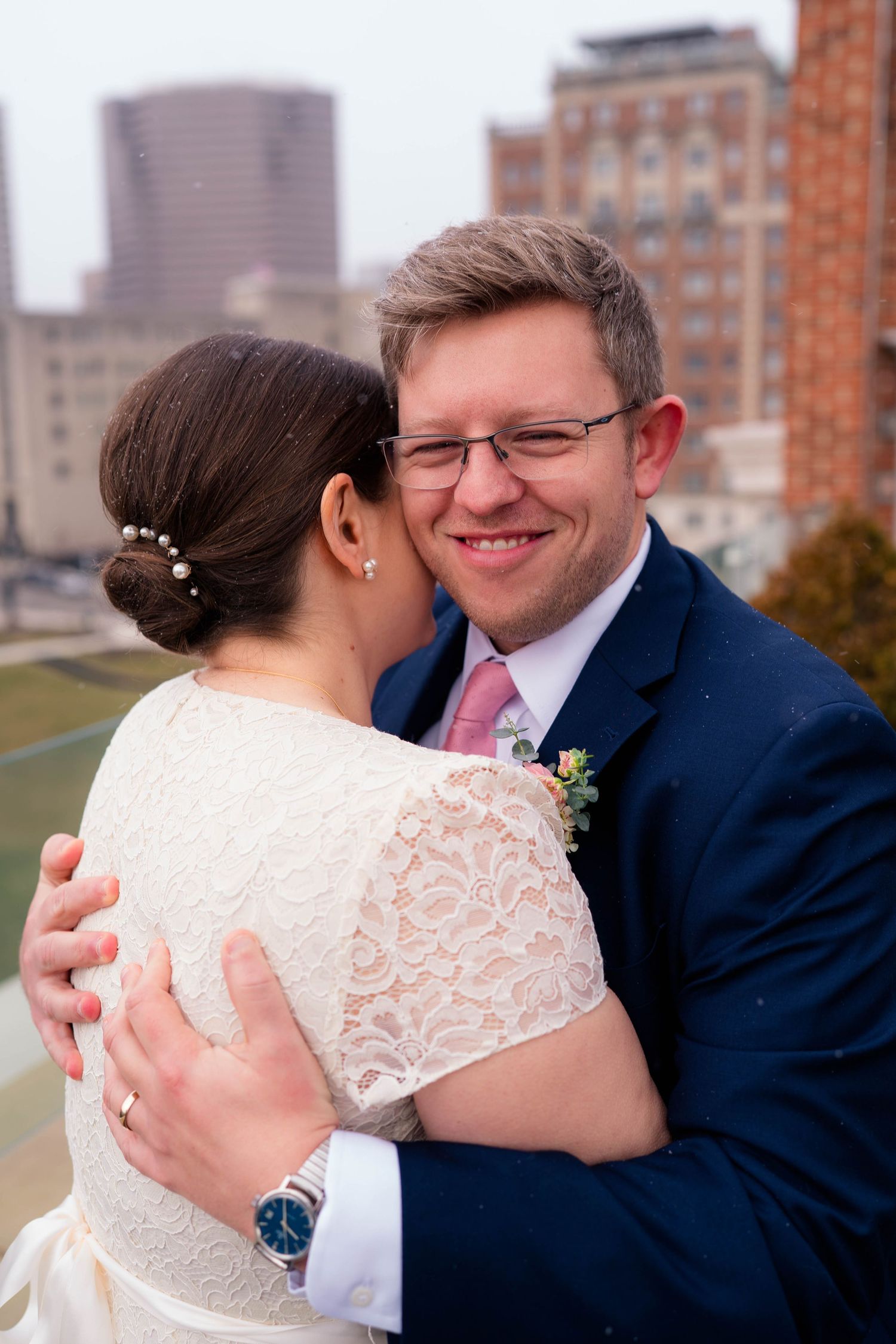 Groom smiling joyfully while embracing bride in white lace dress on rooftop with city backdrop.