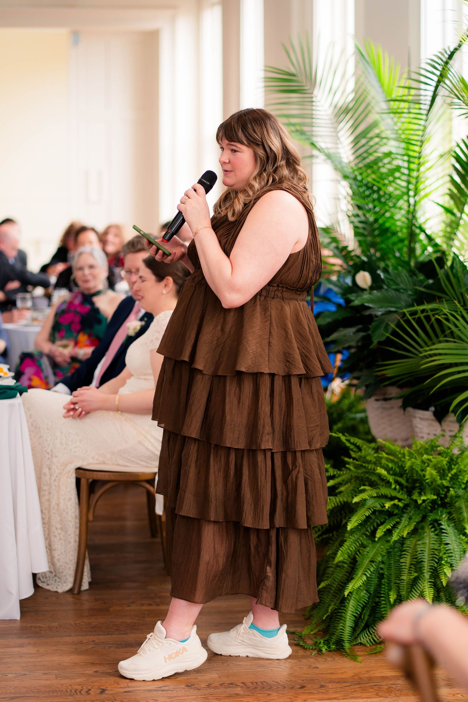 Wedding guest in brown tiered dress and white sneakers delivering a speech with a microphone.