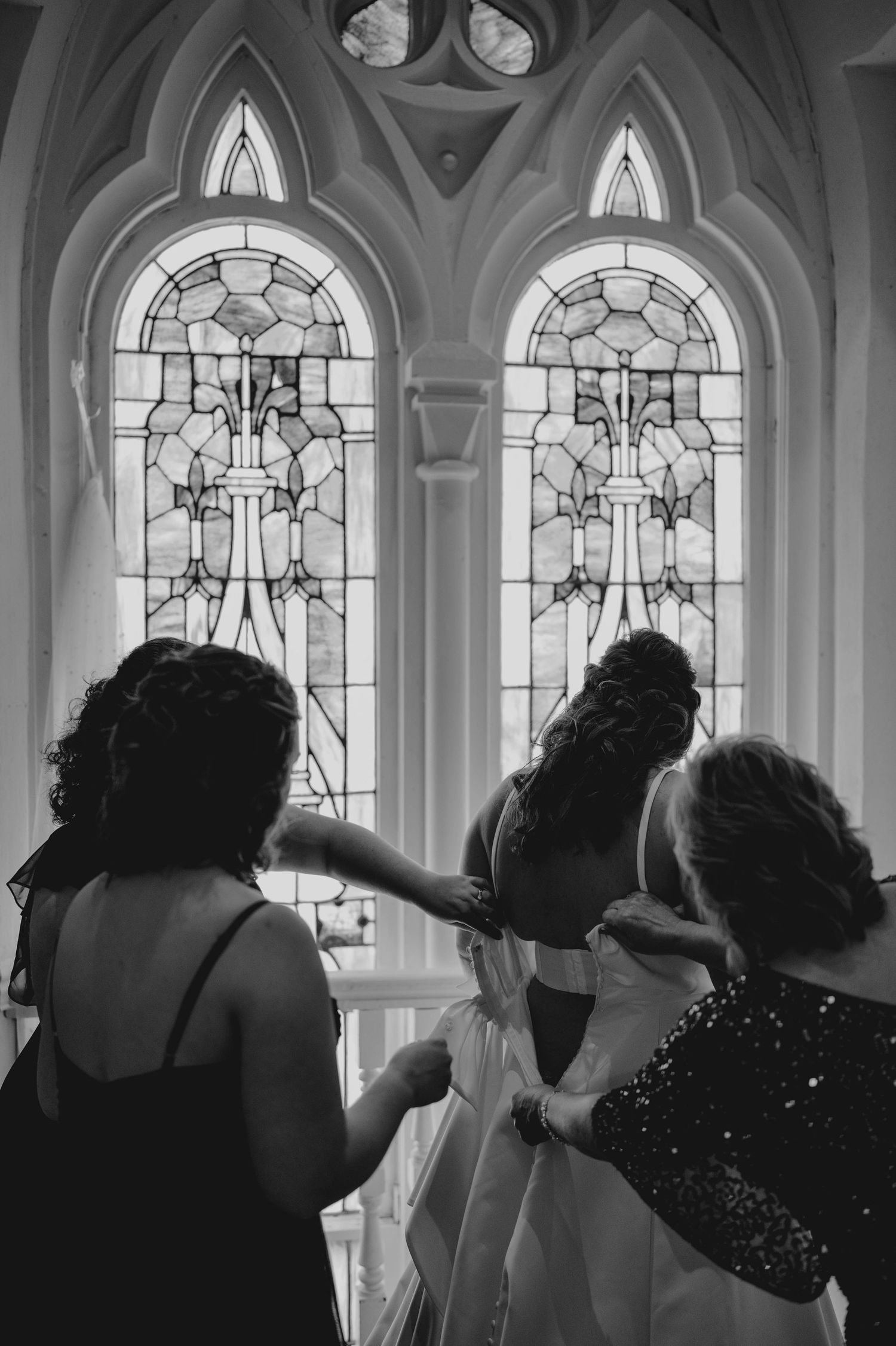 Black and white photo of bridesmaids helping bride with gown before stunning stained glass church windows.