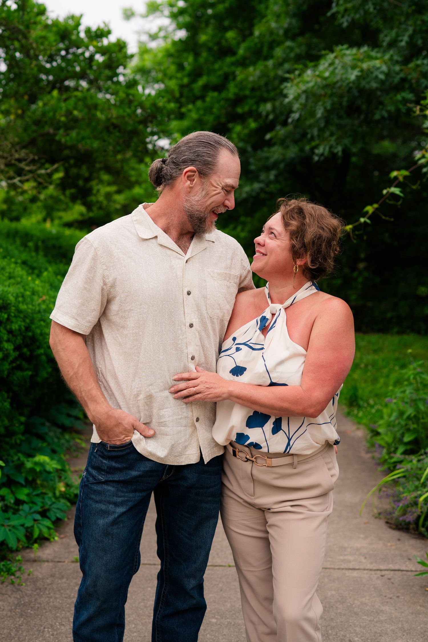 A middle-aged couple smiles at each other on a garden path surrounded by lush green trees.