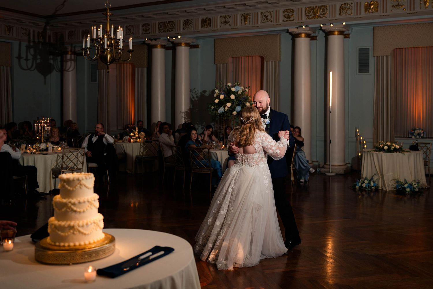 Bride and groom share first dance in elegant ballroom with chandelier, floral centerpieces, and wedding cake nearby.