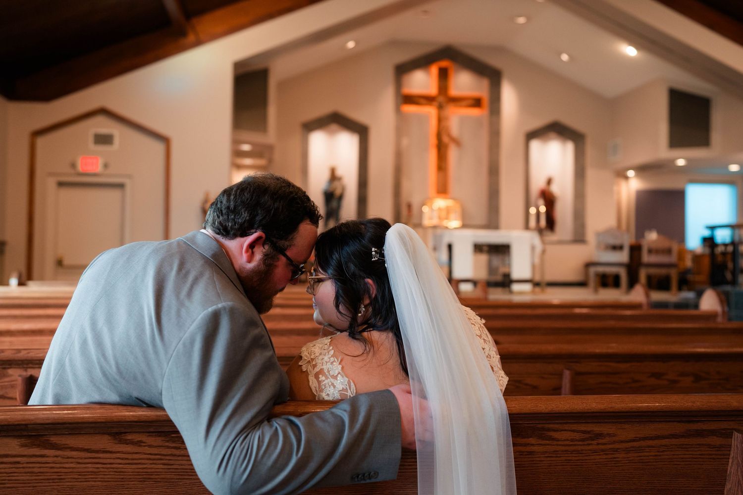 Bride and groom share a quiet prayer together kneeling at church pew before wooden cross.