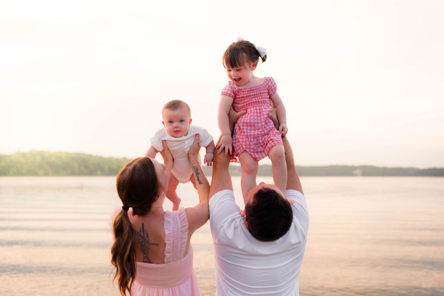 Joyful parents lifting two young babies in the air on a sunny beach with calm water backdrop.