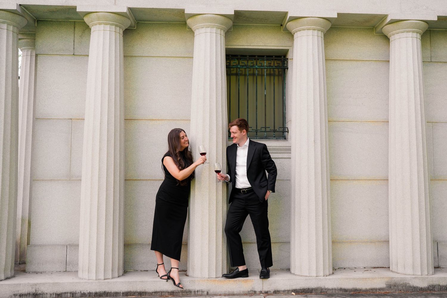 Couple in black formal attire laughing and holding wine glasses beside tall white columns outdoors.