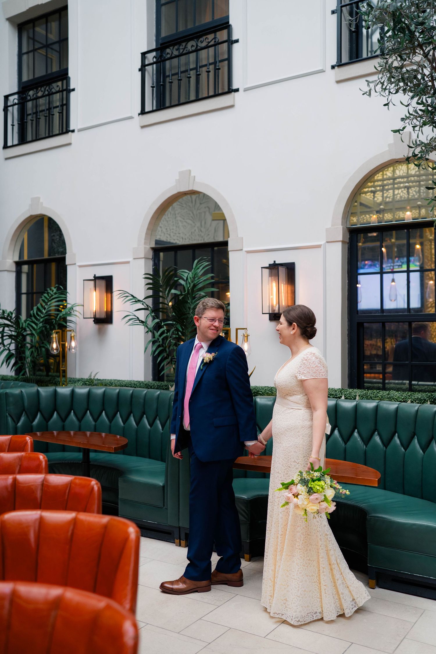Bride in lace gown and groom in pink tie hold hands in elegant hotel courtyard setting.
