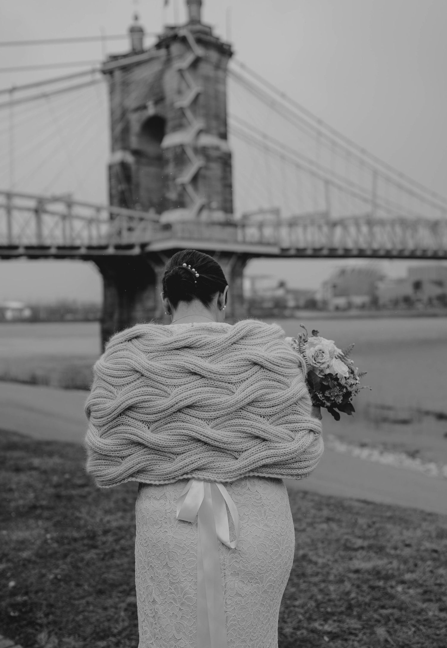 Black and white photo of bride in cable-knit wrap holding bouquet near historic suspension bridge.