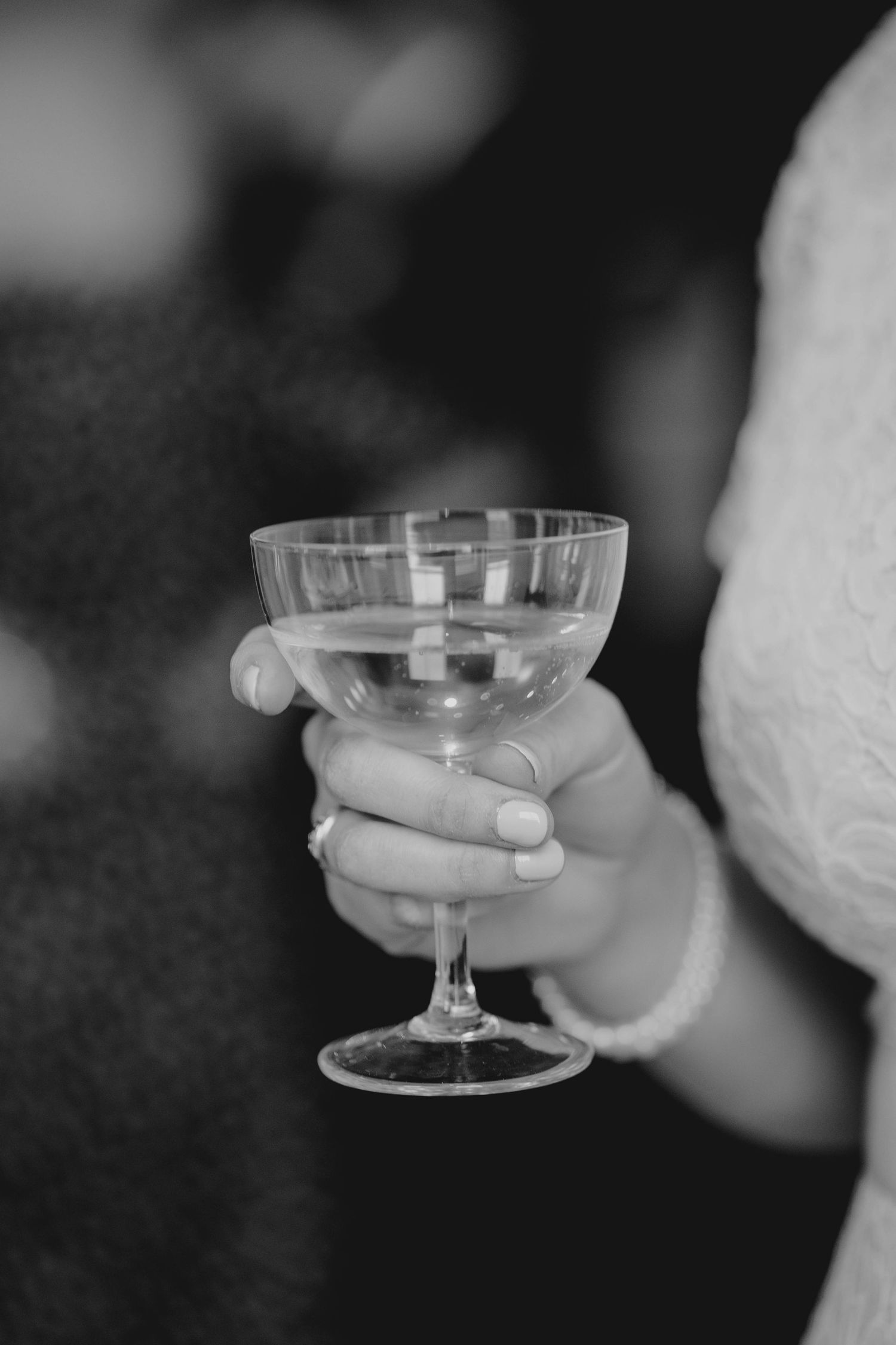 Black and white close-up of bride holding elegant champagne coupe glass with pearl bracelet.