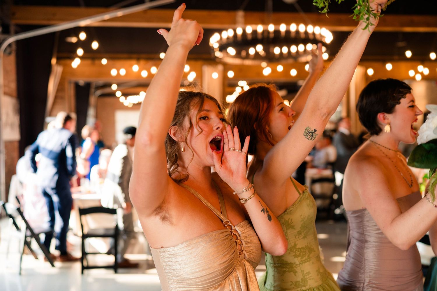 Excited bridesmaids celebrating with arms raised on wedding reception dance floor with string lights.