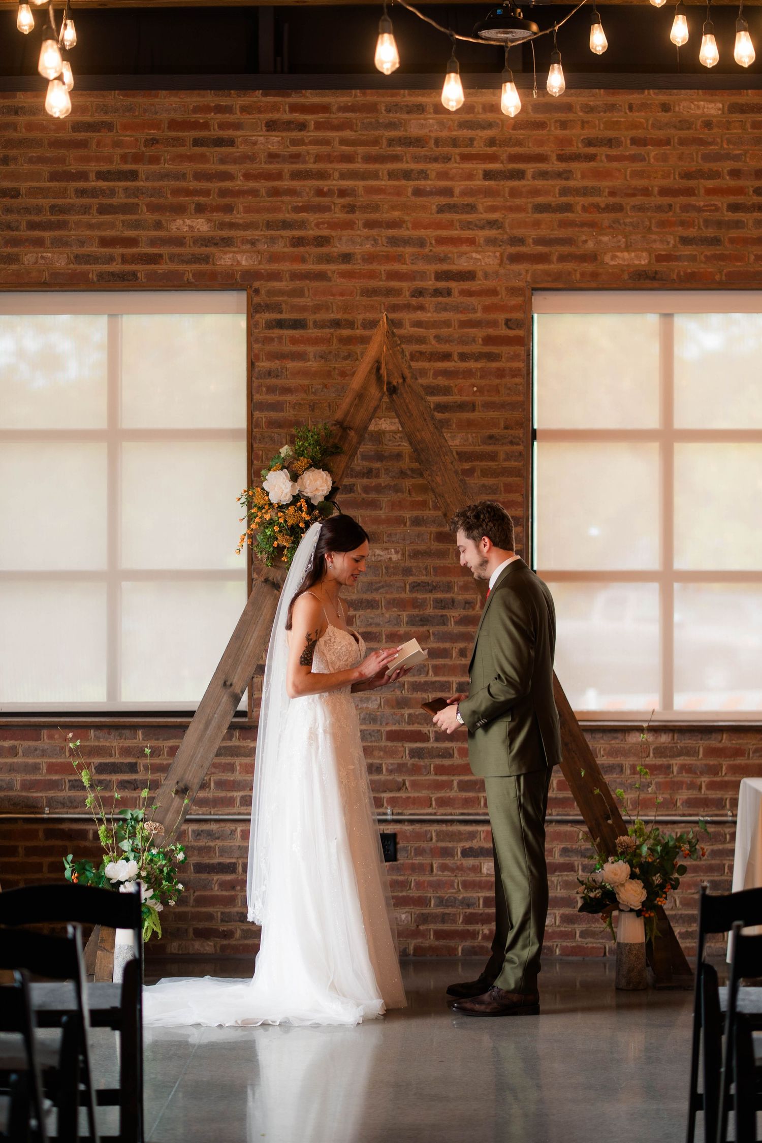 Bride and groom exchanging vows at wooden triangle arch altar in rustic brick wedding venue.