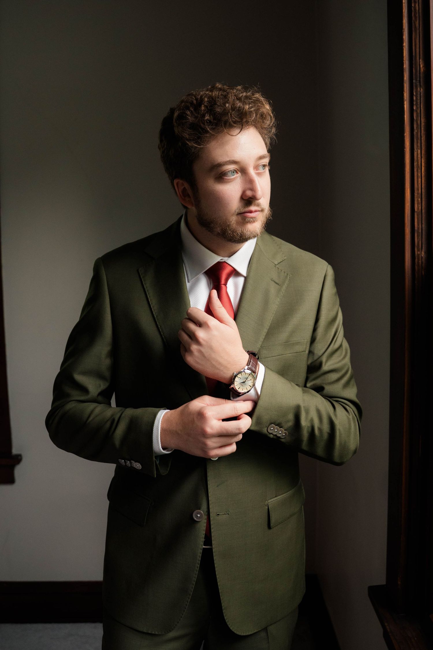 Curly-haired groom in olive green suit and red tie adjusting collar near window.
