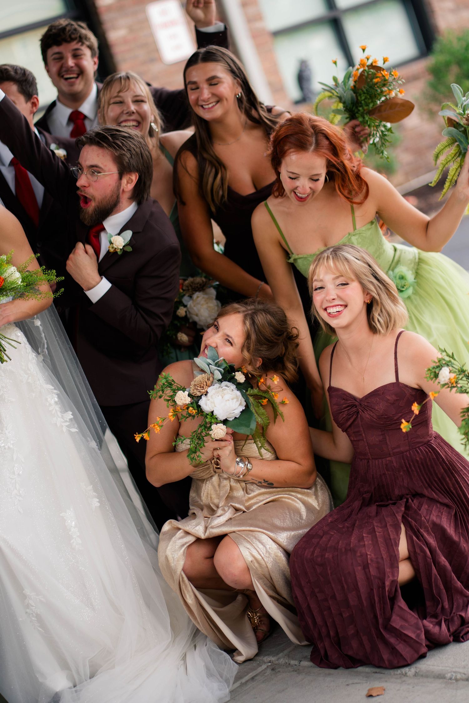 Wedding party laughs and celebrates together on steps, bridesmaids in mixed earth-tone dresses.