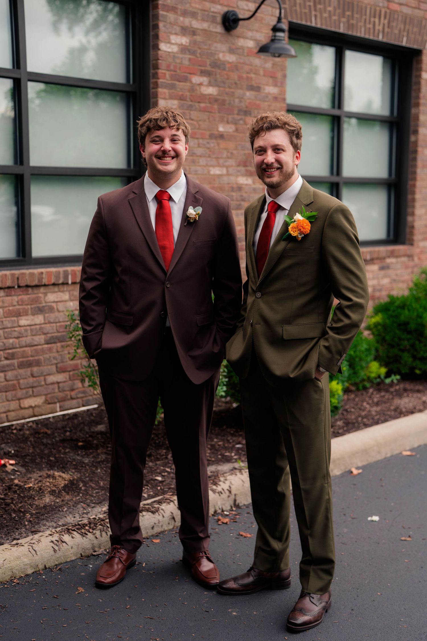 Groom in olive suit and groomsman in brown suit smile wearing orange floral boutonnieres outside brick venue.
