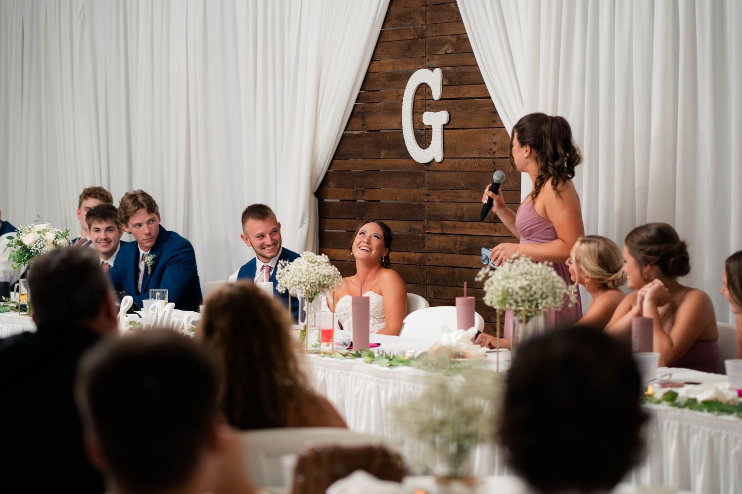 Wedding reception speech by bridesmaid at elegant venue with wooden backdrop, white drapes, and floral arrangements at head table.