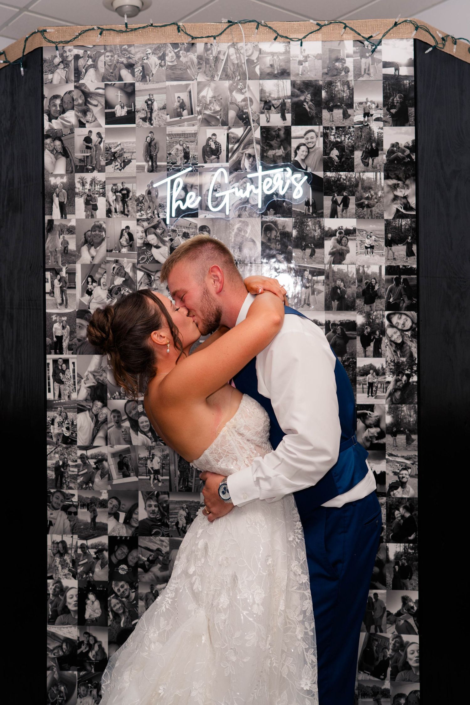 Bride and groom share a romantic kiss in front of a black and white photo collage wall with a neon sign reading The Gunters.