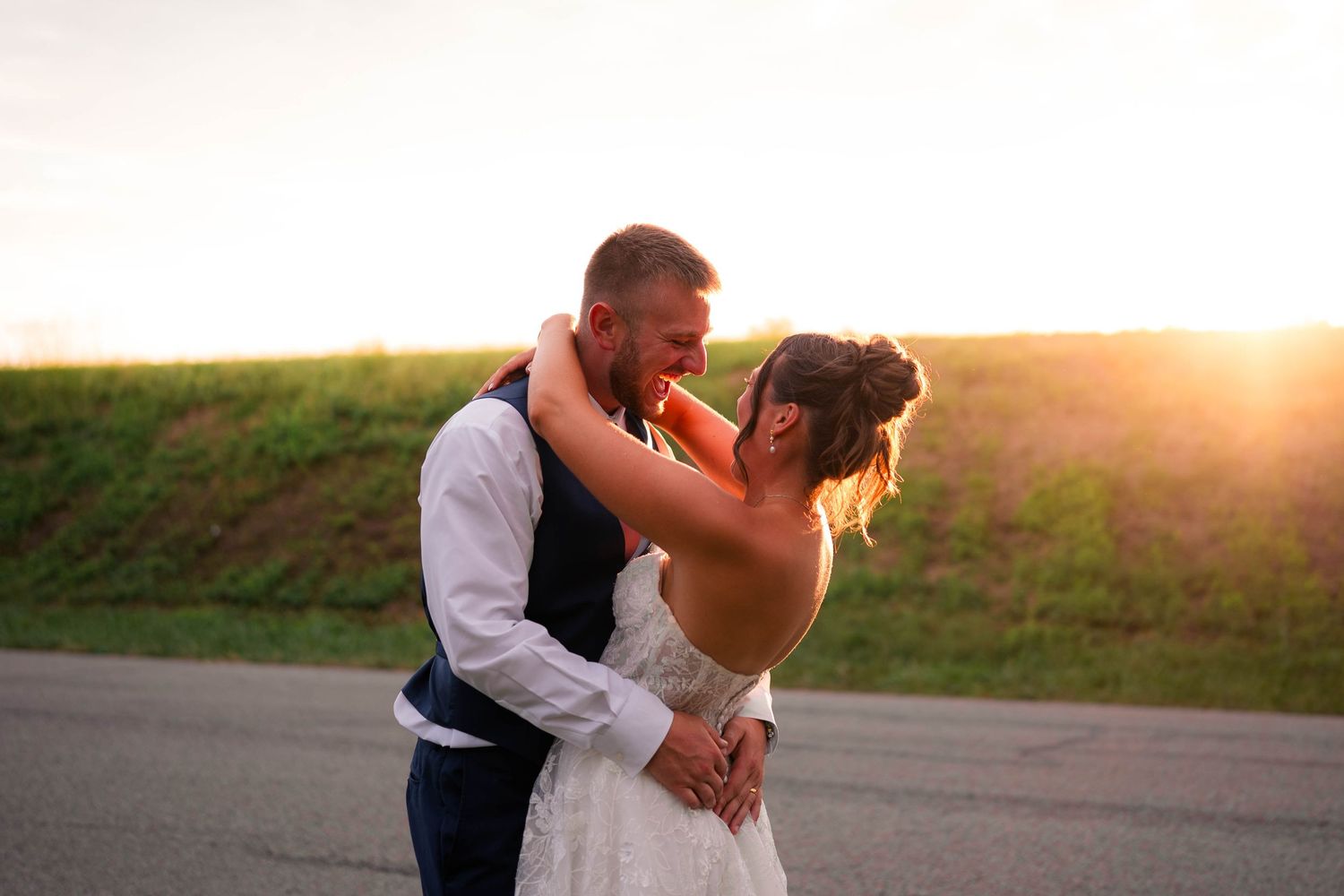 Bride and groom embracing at sunset on a country road, golden light glowing behind them.
