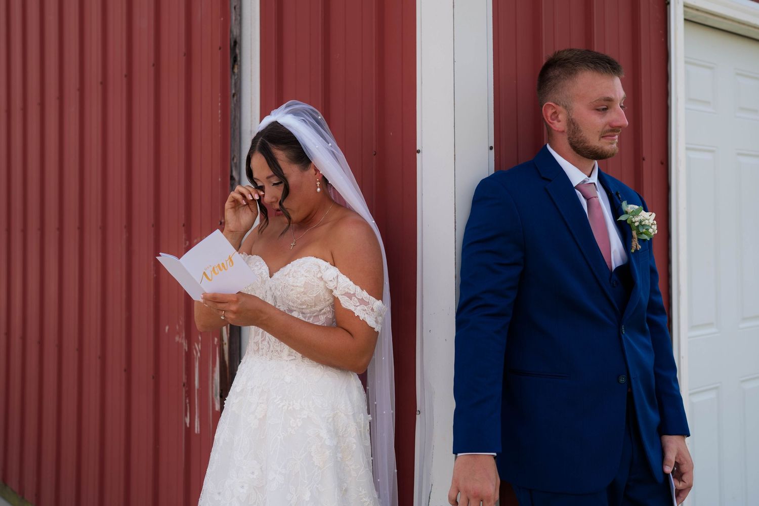 Bride emotional reading vows outside red barn while groom stands with back turned during first look.