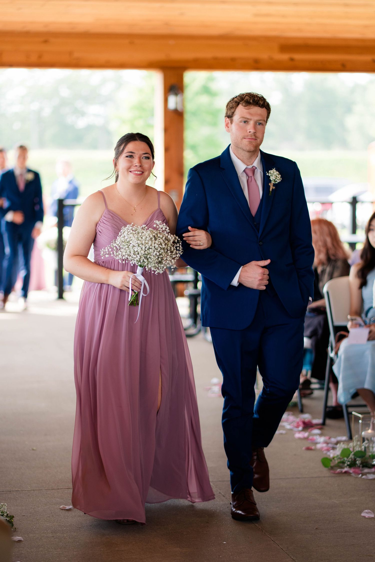 Bridesmaid in dusty rose gown walks down aisle with groomsman in navy suit during ceremony.