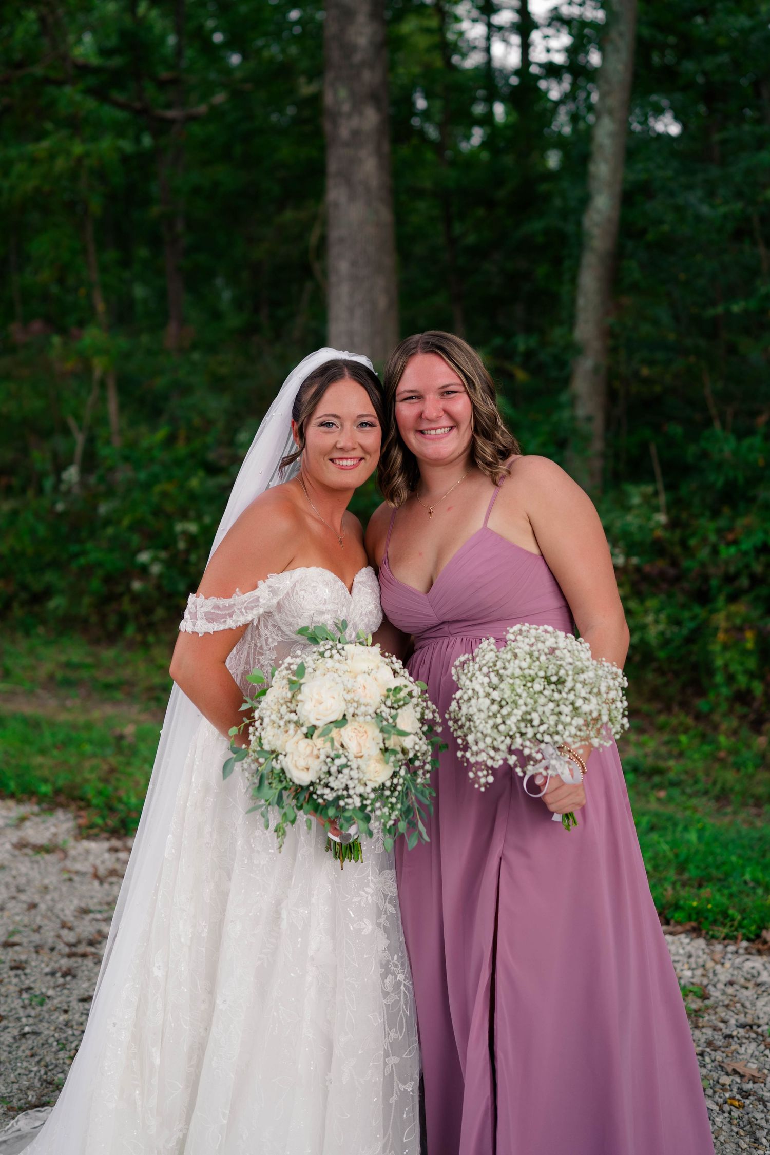 Smiling bride in lace off-shoulder gown and bridesmaid in mauve dress holding white bouquets outdoors at wedding.