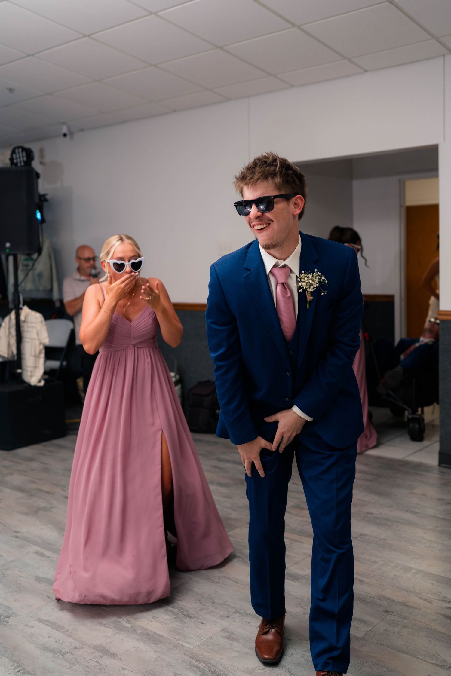 Groomsman and bridesmaid in mauve dress wearing fun sunglasses during lively wedding reception.