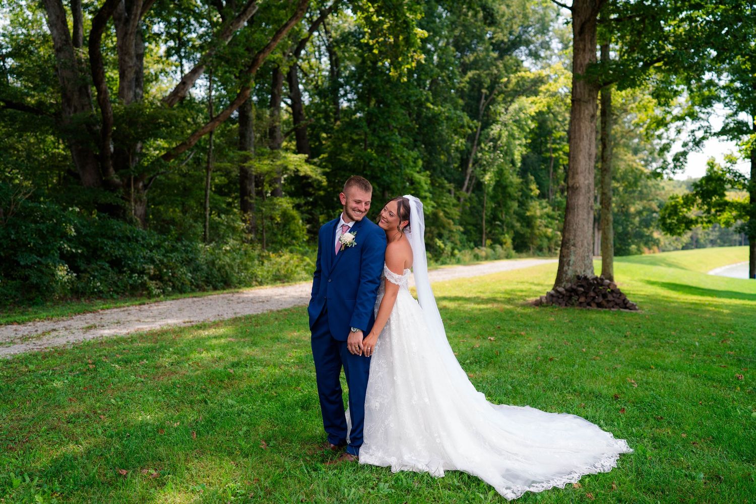 Bride and groom embrace while holding hands on a lush green lawn surrounded by tall trees.