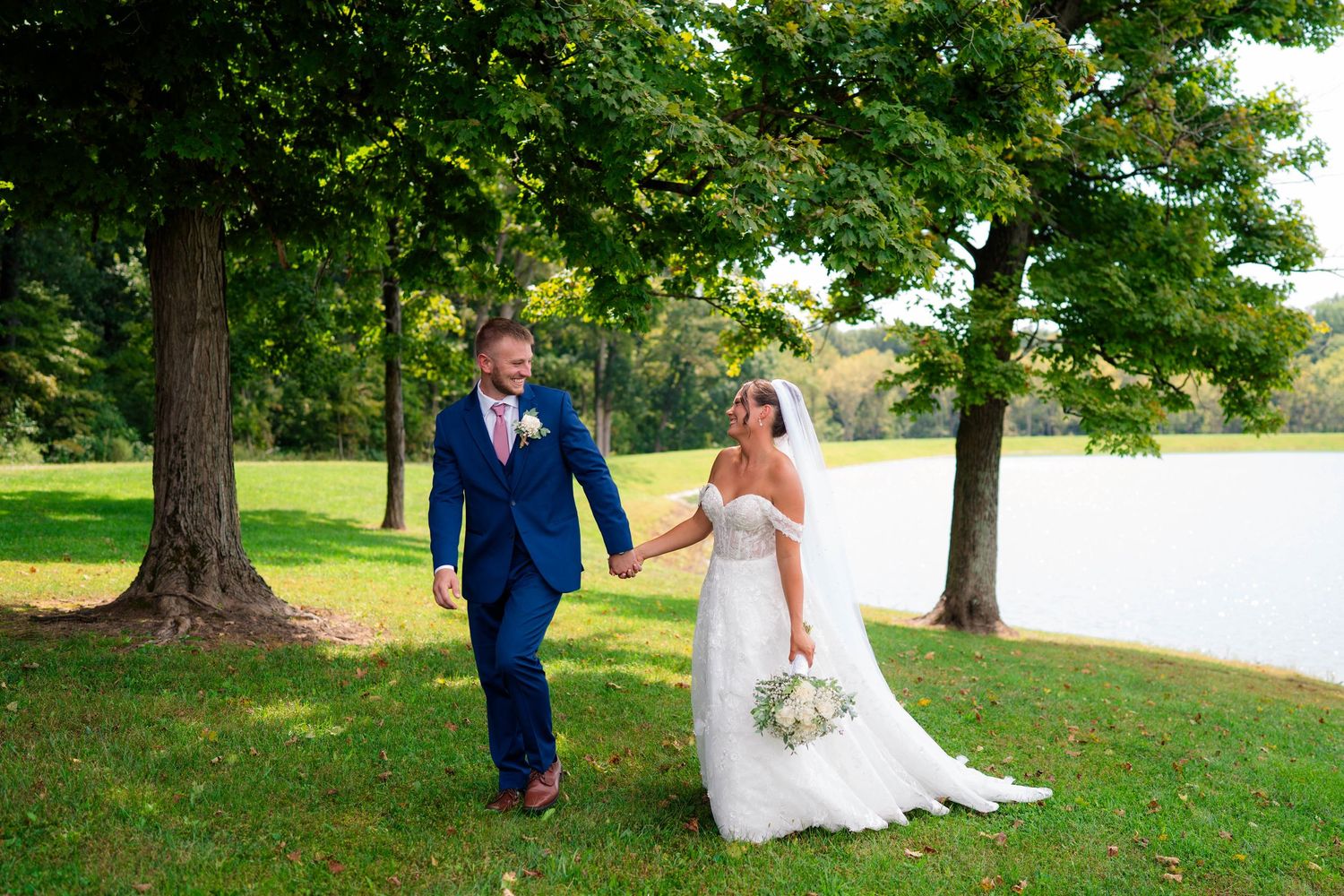 Bride and groom walk hand in hand across a green lawn with a serene pond behind them.