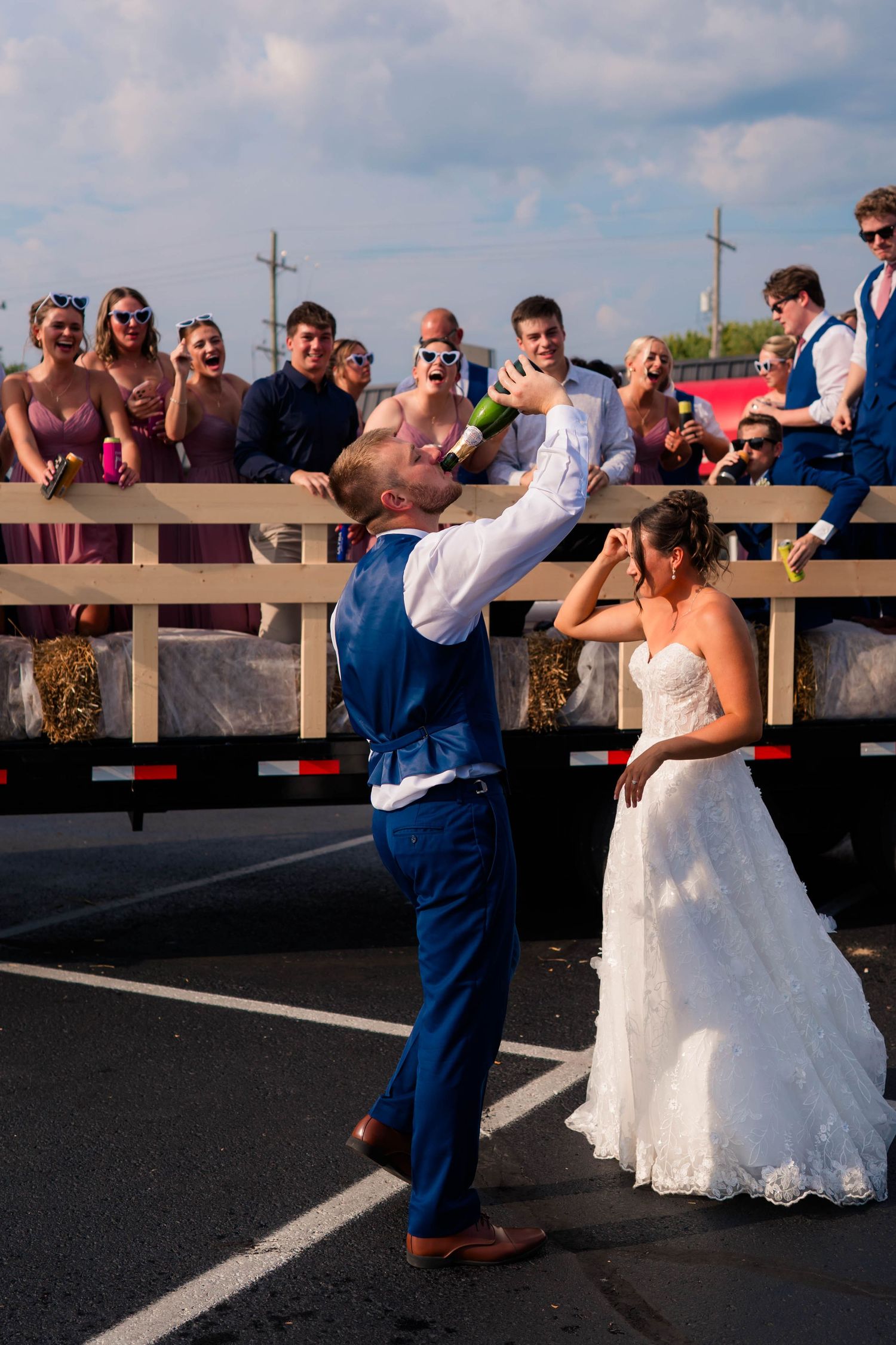 Groom catches a tossed bottle while bride watches, with wedding guests cheering from a flatbed truck.