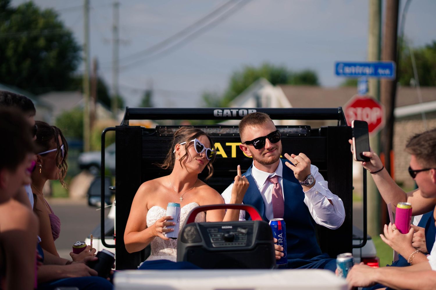 Bride and groom wear sunglasses and smile while riding in a white Gator vehicle after wedding ceremony.