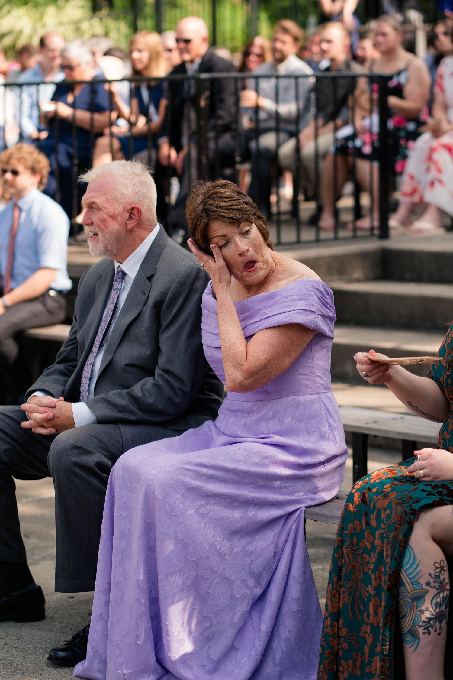 Emotional woman in lavender gown wipes tears at outdoor wedding ceremony seated beside man in gray suit.