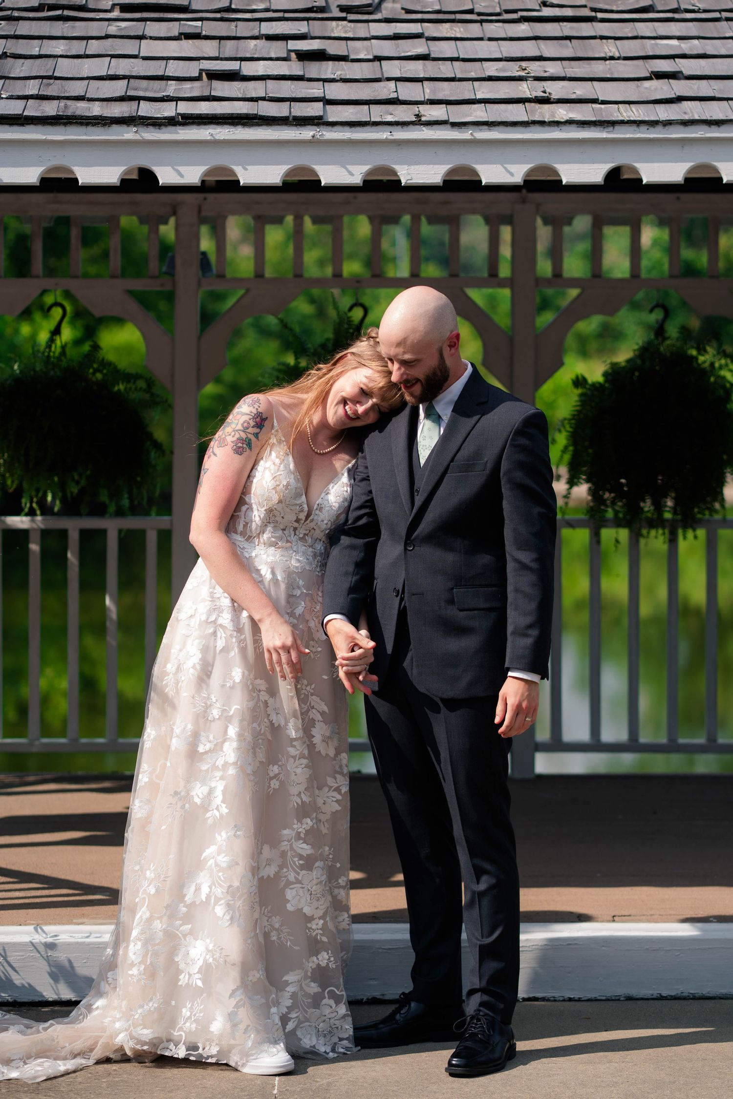 Bride in lace gown leans on bald groom while holding hands in front of white gazebo on sunny wedding day.