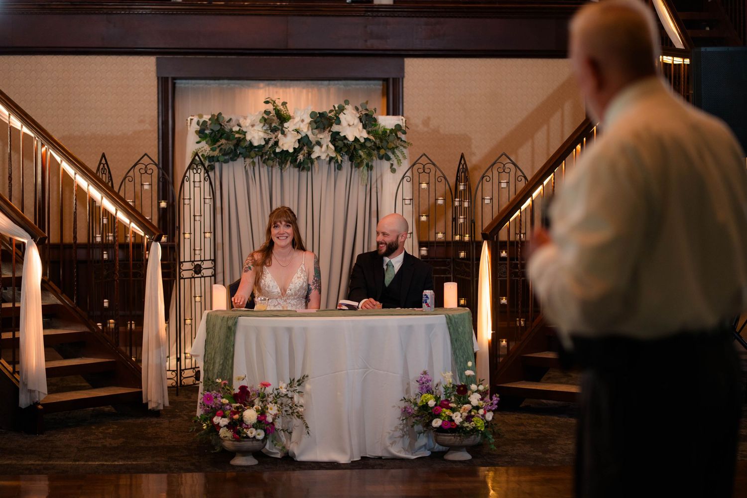 Bride and groom at decorated sweetheart table with greenery arch and candles as guest gives toast at wedding reception.