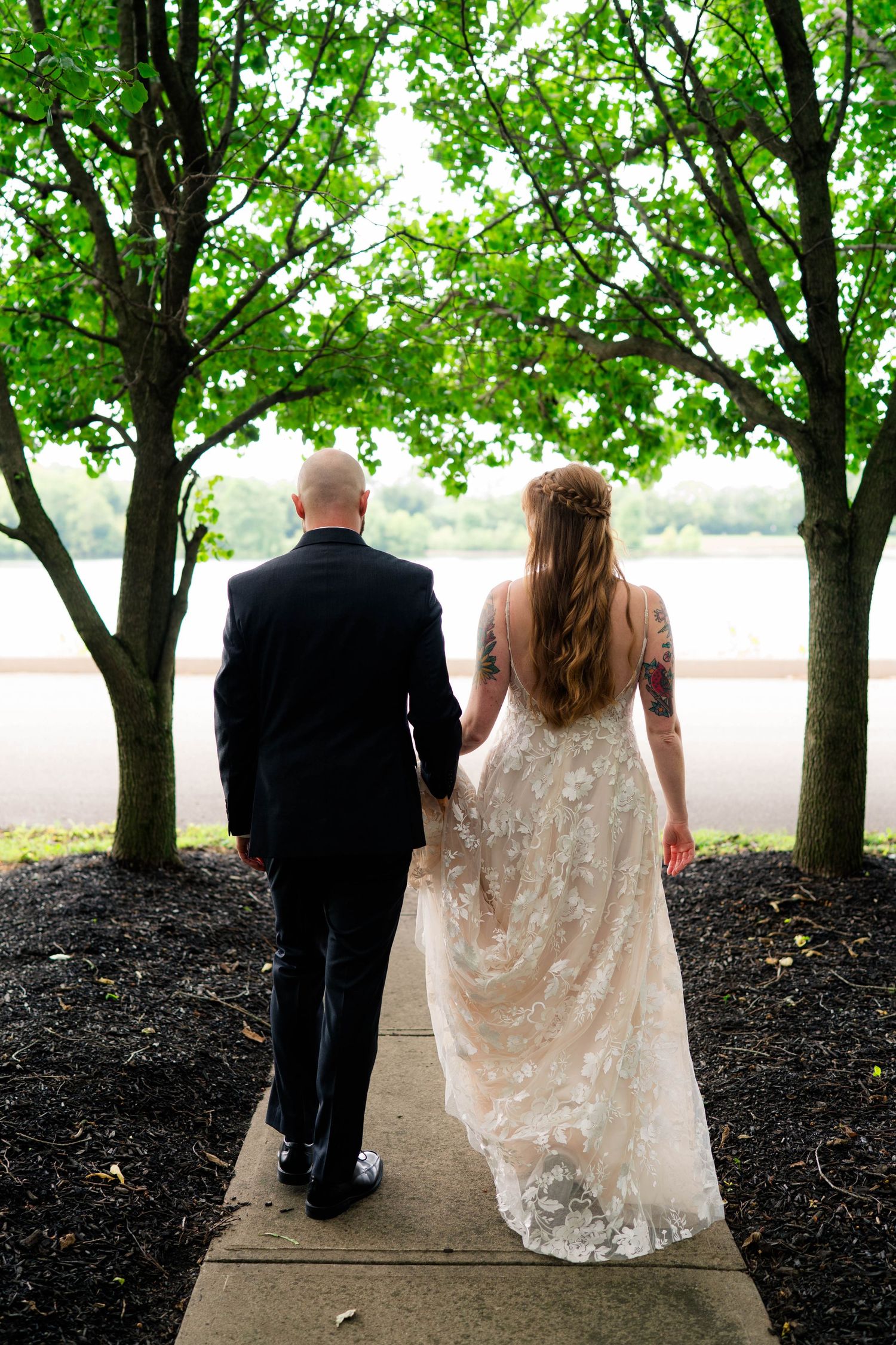 Bride and groom walking hand in hand along a tree-lined path toward a serene waterfront on their wedding day.