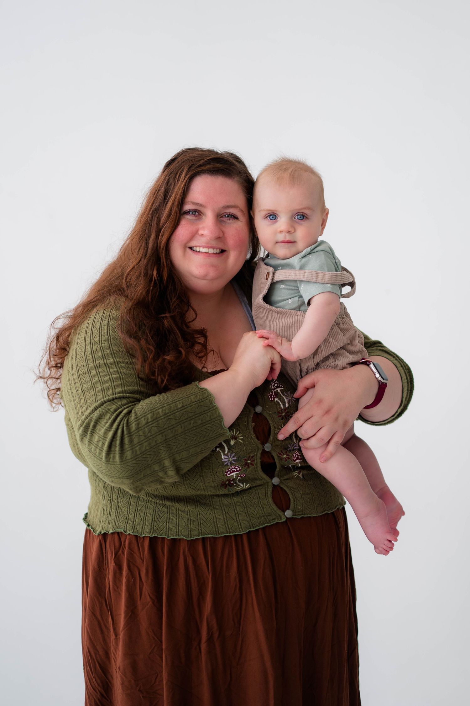 Smiling woman in green top holding a blue-eyed baby in brown outfit against white background.