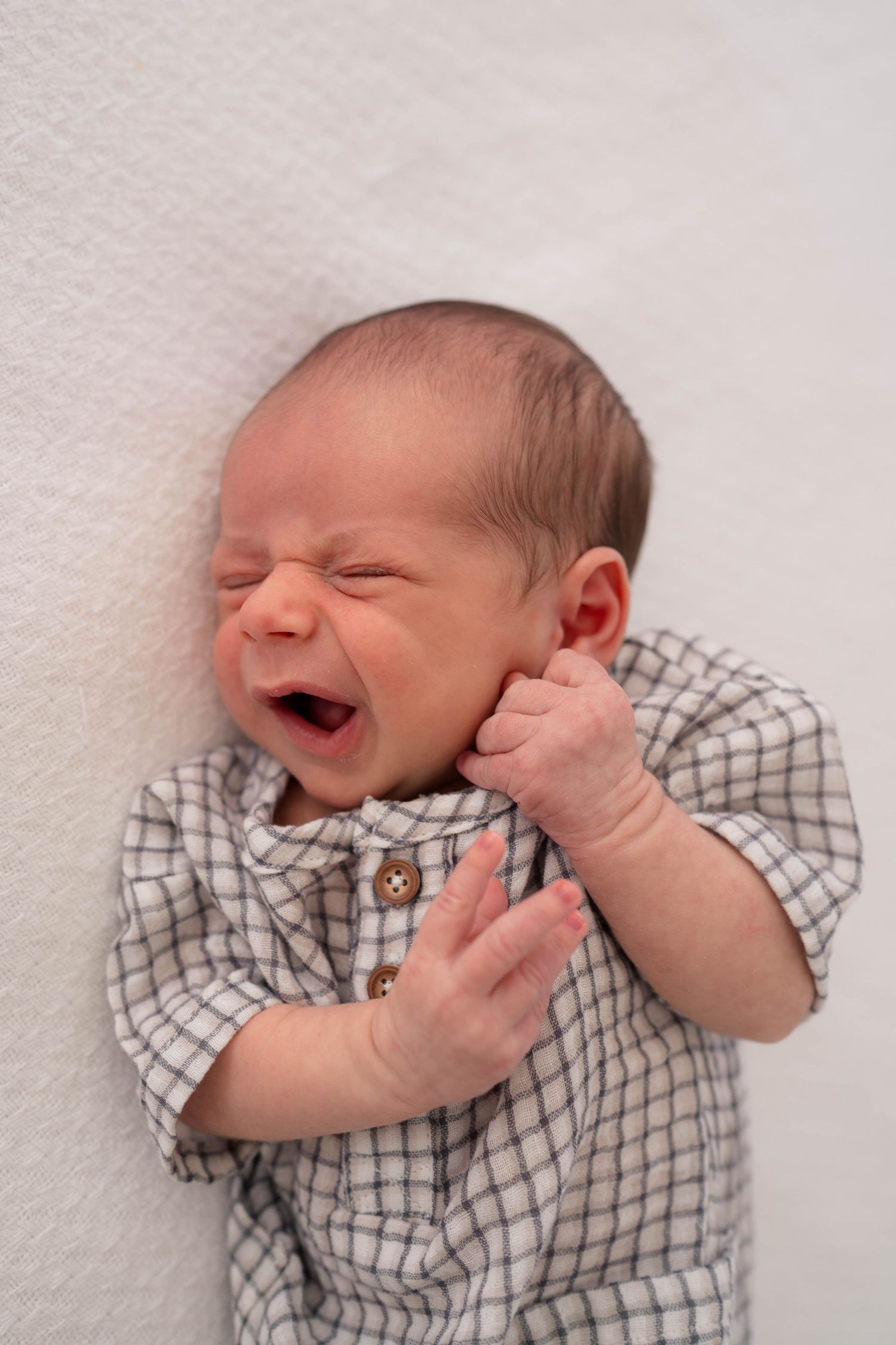 Newborn baby yawning in a gray plaid onesie, lying on a white blanket.