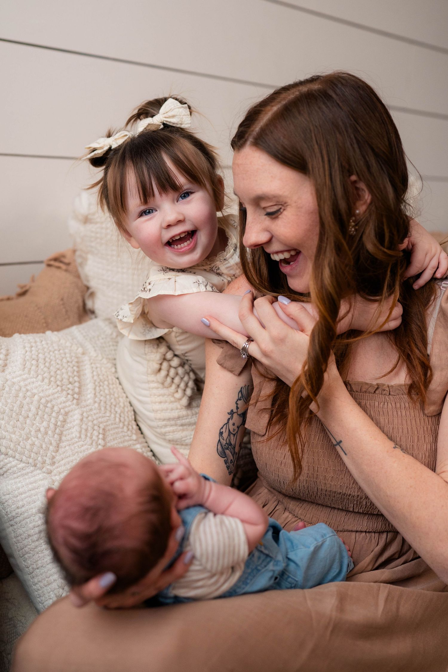 Joyful mother laughing with excited toddler daughter in cream dress while holding newborn baby.